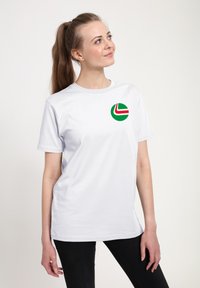 Bílý tričko s krátkým rukávem a kulatým zeleným logem obsahujícím červený a bílý geometrický design na levé části hrudi. Měkký materiál, volný střih.