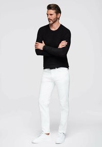 Schwarzes, strukturiertes Langarmshirt kombiniert mit weißen Jeans und weißen Turnschuhen. Das Model steht mit verschränkten Armen vor einem hellgrauen Hintergrund.
