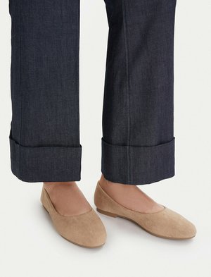 Persona con pantalones oscuros remangados y zapatos planos de ante beige de pie sobre una superficie clara.