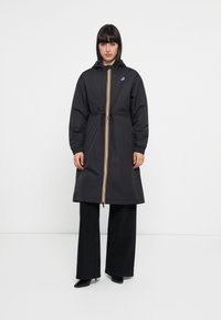 Black hooded raincoat with a yellow and red zipper, cinched waist, and elastic cuffs. Styled with wide-legged black pants.