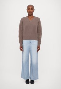 Brauner, gestrickter V-Ausschnitt-Pullover kombiniert mit weit geschnittenen, hellblauen Jeans und schwarzen Schuhen, die eine lässige Passform und strukturierte Stofflichkeit zur Schau stellen.