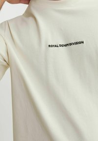 Kreminės spalvos trumpa rankovė t-shirt, pagaminta iš švelnios medvilnės, su juodu siuvinėtiniu tekstu "ROYAL DENIM DIVISION" viršutinėje nugaros dalyje.
