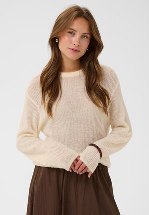 Jeune femme aux longs cheveux bruns portant un pull en maille crème et une jupe marron, debout, les mains jointes devant un fond uni.