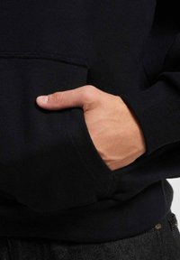 Crna dukserica s kenguruvim džepom. Tkanina je teksturirana i mekana, s rebrastim manžetama. Ruka se ležerno oslanja na džep.