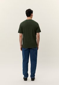 Olivově zelené bavlněné tričko s kulatým výstřihem, krátkými rukávy a volným střihem, v kombinaci s modrými džínami a černými botami.