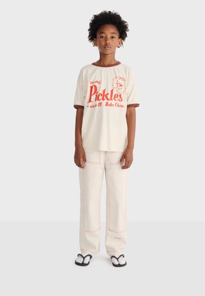 Enfant aux cheveux bouclés portant un T-shirt blanc avec un motif de chien rouge et du texte, un pantalon blanc avec des coutures rouges, et des sandales noires et blanches.