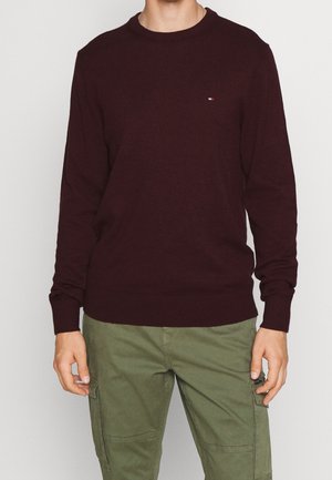 Homme portant un pull manches longues bordeaux foncé avec un petit logo sur la poitrine, et un pantalon cargo vert olive, debout devant un fond uni.