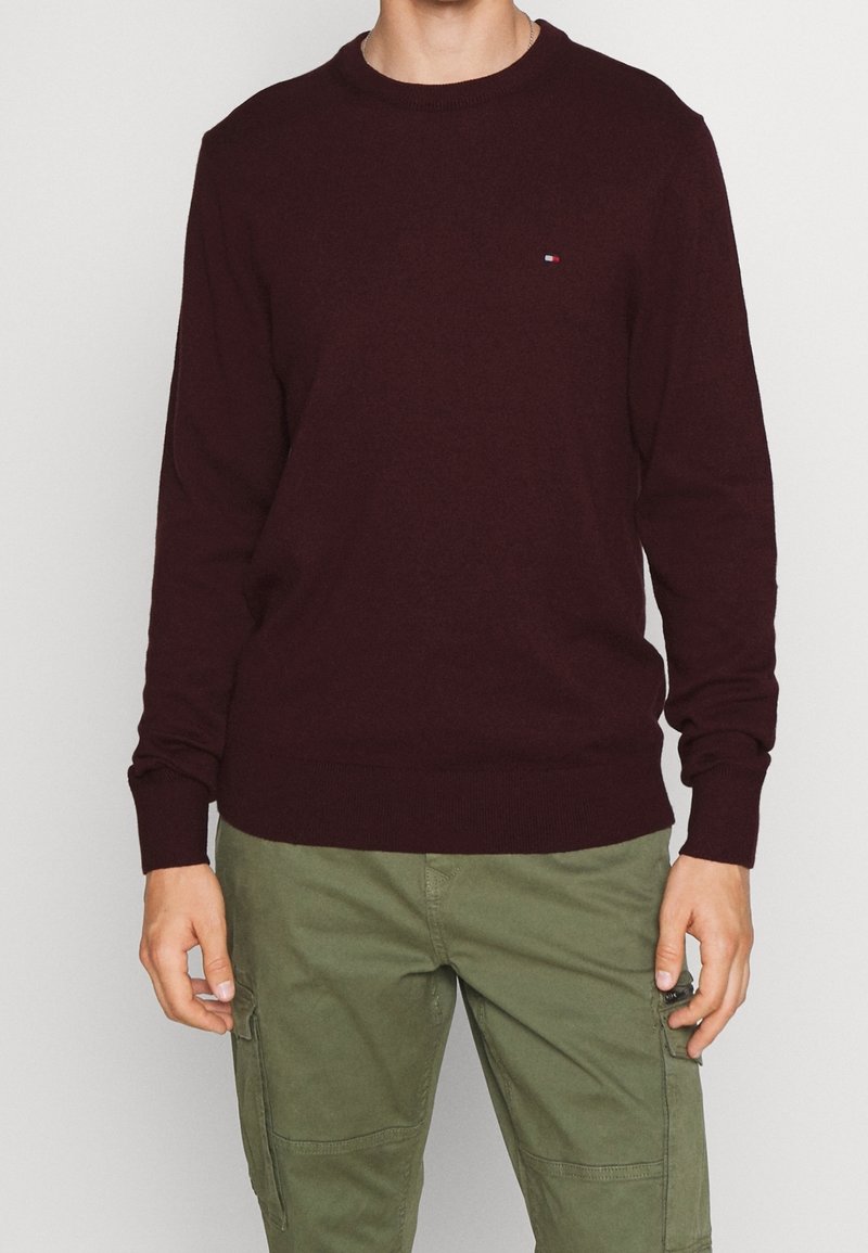 Homme portant un pull manches longues bordeaux foncé avec un petit logo sur la poitrine, et un pantalon cargo vert olive, debout devant un fond uni.