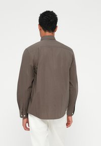 Camisa marrón de mangas largas con rayas verticales, cuello abotonado y puños abotonados, presentada desde la parte posterior en un modelo que lleva pantalones blancos.
