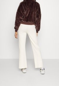 Brązowa kurtka z sztucznego futra z wysokim kołnierzem i elastycznymi mankietami, zestawiona z białymi spodniami dzwonami i sportowymi białymi sneakersami.