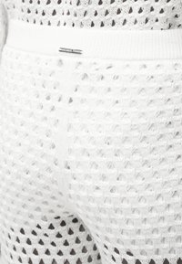 Κοντινό πλάνο λευκού πλεκτού παντελονιού με μοτίβο δίχτυ και μικρή μεταλλική ετικέτα με την επωνυμία "Michael Kors" στη μέση.