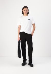 Vit t-shirt med kort ärm och en liten logotyp, svarta byxor och svarta skor. Personen håller en svart tote-väska. Enkelt design och neutrala färger.