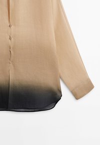 Chemise beige à manches longues avec de petits boutons sur le devant, présentant un dégradé allant du beige au noir au niveau de l'ourlet inférieur et des poignets.