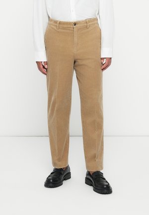 Beige corduroy pants worn with a white shirt and black leather loafers, standing against a plain white background.