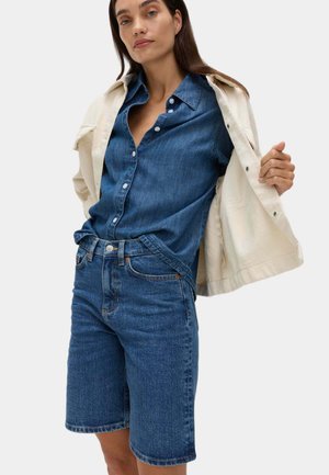 Kvinde iført blå denimskjorte og shorts, holder en beige jakke åben mod en ensfarvet baggrund.