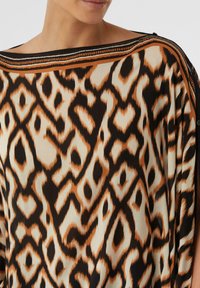 Luźna bluzka z lekkiego materiału, ozdobiona wyraźnym zwierzęcym wzorem w kolorach czarnym, beżowym i pomarańczowym, z szerokim dekoltem i dekoracyjnym wykończeniem.