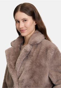 Ung kvinde med langt brunt hår iført en blød, lysebrun faux fur-frakke, der smiler svagt foran en ensfarvet baggrund.