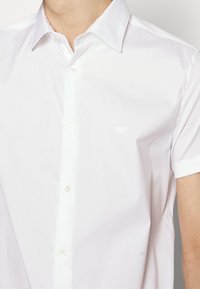 Muž v bílé košili s krátkým rukávem a špičatým límečkem, na hrudi má diskrétní vyšívané logo.