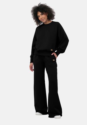 Kobieta o kręconych włosach, ubrana w luźną czarną bluzę i szerokie spodnie, stojąca z jedną ręką w kieszeni na jednolitym tle.