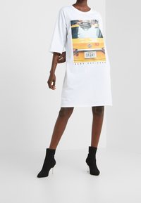 Weißes, übergroßes T-Shirt-Kleid mit einem Grafikdruck eines gelben Taxis, kurzen Ärmeln und einem runden Ausschnitt. Mit dem Text "DKNY" und "EST. 1989".
