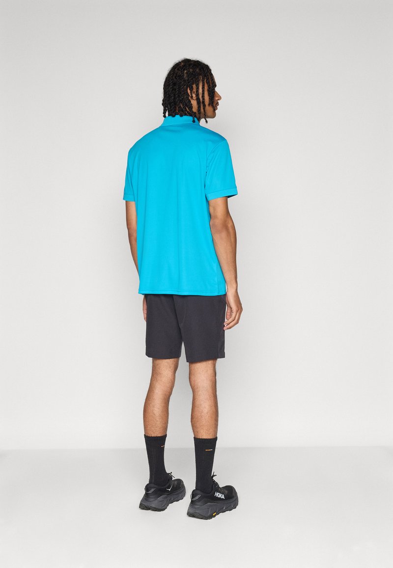 Türkisfarbiges Polo-Shirt mit kurzen Ärmeln, kombiniert mit schwarzen Shorts. Modell trägt schwarze Socken und Schuhe und steht mit dem Rücken zur Kamera.