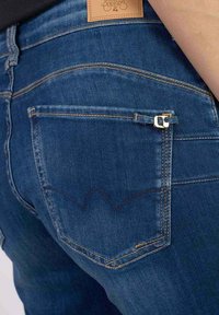 Jeans en denim bleu foncé avec une poche arrière dotée d'une petite fermeture éclair dorée et d'un motif brodé distinctif en forme de vague au-dessus de la poche.