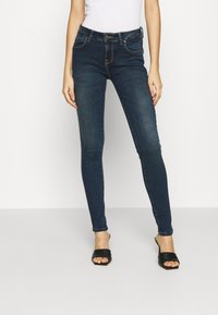 Vaqueros oscuros ajustados de tiro medio, con cinco bolsillos y un sutil efecto desgastado, combinados con sandalias negras tipo slip-on.