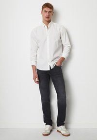 Witte button-up shirt, donkergrijze jeans en witte sneakers met beige accenten. Model staat met één hand in de zak tegen een effen achtergrond.