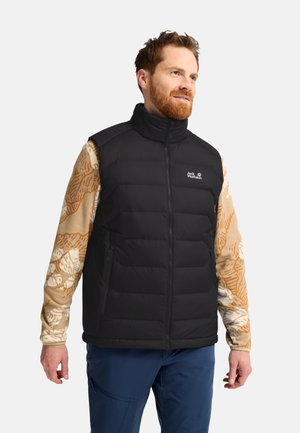 Mann trägt eine schwarze, gesteppte ärmellose Weste über einem beige gemusterten Langarmhemd und blauen Hosen und schaut zur Seite.