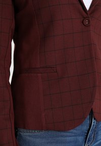 Burgundský kostkovaný blejzr s jedním knoflíkem, s rovným kapsou, nošený přes bílou košili a modré džíny.