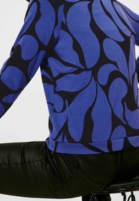 Modrá mikina se abstraktními tmavě modrými listovými vzory, vyrobená z měkké látky, spárovaná s černými kalhotami z umělé kůže.