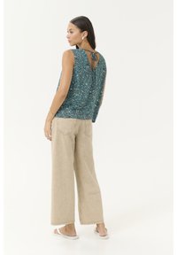 Frau in ärmellosem, türkisfarbenem, gemustertem Oberteil mit Rückenknoten, beigen weiten Hosen und weißen Sandalen, die vor einem weißen Hintergrund steht.