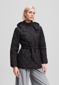 Veste matelassée noire avec une taille ceinturée, capuche réglable et boutons-pression, dotée d'une finition lisse et de deux poches avant.