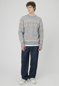 Pull gris à motifs géométriques blancs et jaunes, associé à un pantalon en denim bleu foncé et des baskets blanches.