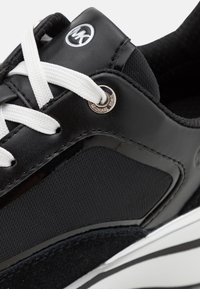 Schwarzer Sneaker mit einer Kombination aus glattem Leder und strukturiertem Stoff, weißen Schnürsenkeln und einem Metall-Logo-Akzent an der Seite.