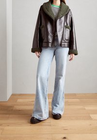 Veste en cuir marron avec col en fausse fourrure verte et poches, associée à un jean évasé bleu clair et des chaussures compensées marron foncé.