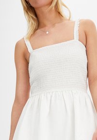 Vit klänning med smockad liv, tunna volangaxlar och en utsvängd kjol. Tillverkad av lätt tyg med en mjuk textur.