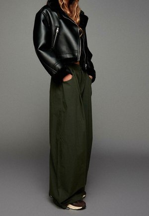 Persona con chaqueta de cuero negra recortada con borde de piel, pantalones anchos verde oscuro con las manos en los bolsillos y zapatillas oscuras sobre fondo gris.