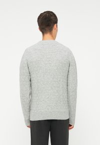 Pull en maille grise avec motif en losanges, poignets et bas côtelés, design col rond. Porté par-dessus un pantalon sombre, vu de dos.