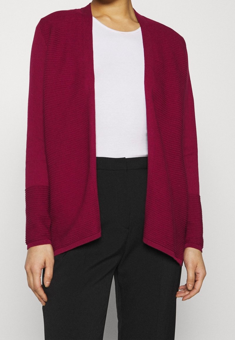 Femme portant un cardigan texturé, ouvert sur le devant, de couleur rouge foncé, sur une chemise blanche et un pantalon noir, sur un fond uni.