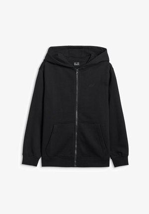Fille aux longs cheveux portant un bandeau blanc, une veste noire zippée, un pantalon noir, une chemise blanche, tenant un petit sac violet, debout sur un fond uni.