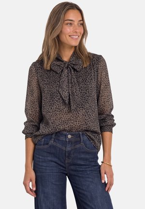 Femme aux cheveux mi-longs porte un chemisier transparent marron foncé à motifs avec un nœud au col et un jean bleu foncé, souriante et regardant vers la droite.