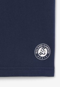 Tela azul marino con un pequeño parche circular con el logo que dice "Roland Garros París" en texto y diseño blancos cerca del borde inferior.