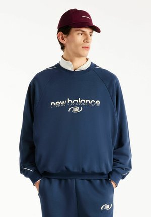 Giovane uomo che indossa felpa e pantaloni New Balance blu navy con logo bianco, abbinati a un berretto New Balance color bordeaux, guarda di lato.
