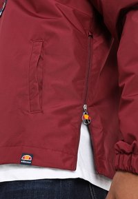 Chaqueta ligera en color burdeos con cierre de cremallera, puños elásticos, bolsillo lateral y un parche de marca con un logotipo en color en el dobladillo.