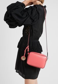 Różowa torba na ramię o teksturowanym wykończeniu, prostokątnym kształcie, złotych akcentach i odpinanym charmsie, noszona z czarną bluzką z koronkowymi detalami.