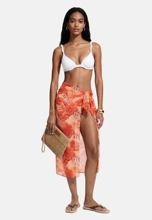 Donna che indossa un top bikini bianco e un pareo floreale arancione annodato alla coscia, con in mano una pochette intrecciata, indossando infradito e un braccialetto d'oro.