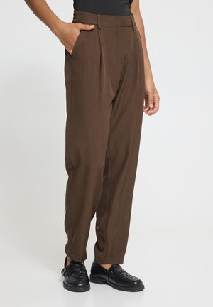 Persoon met een hoge bruine geplooide broek, zwarte loafers en een zwart topje, één hand in de zak, tegen een effen achtergrond.
