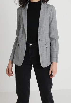 Frau trägt einen grauen karierten Blazer über einem schwarzen Top, kombiniert mit hochtaillierten schwarzen Hosen, und steht vor einem schlichten Hintergrund.