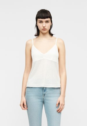 Ung kvinde med kort mørkt hår iført en hvid ærmeløs V-hals top og lyseblå jeans, stående mod en ensfarvet lys baggrund.